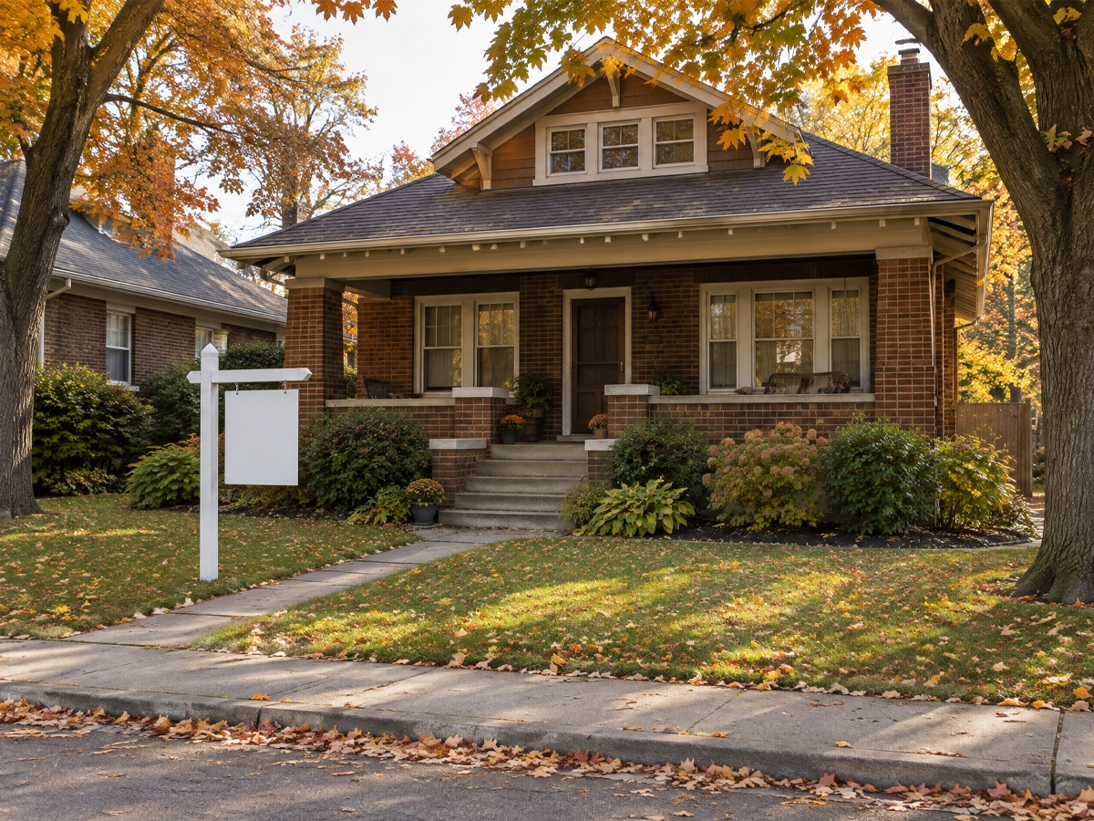 Older brick bungalow with a deep porch and mature maples in autumn, a blank real estate sign on the front lawn