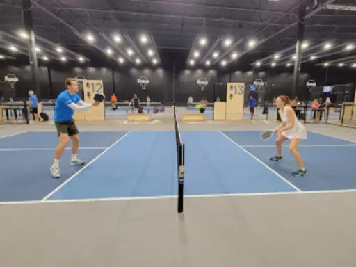 Two people play a game of pickleball indoors while Rhino Rooter workers talk about fixing a sewer nearby.